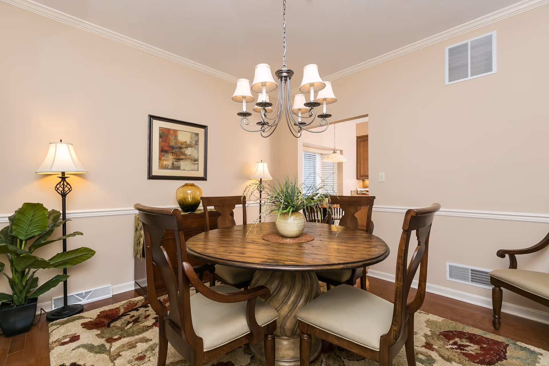 A cozy dining room with a round wooden table surrounded by four cushioned wooden chairs. A decorative plant sits in the center of the table. The room features beige walls with white trim, a patterned area rug, two floor lamps, a framed abstract painting, and a chandelier with six lampshades hanging above the table. A doorway leads to a kitchen area with visible cabinets and a window with blinds.