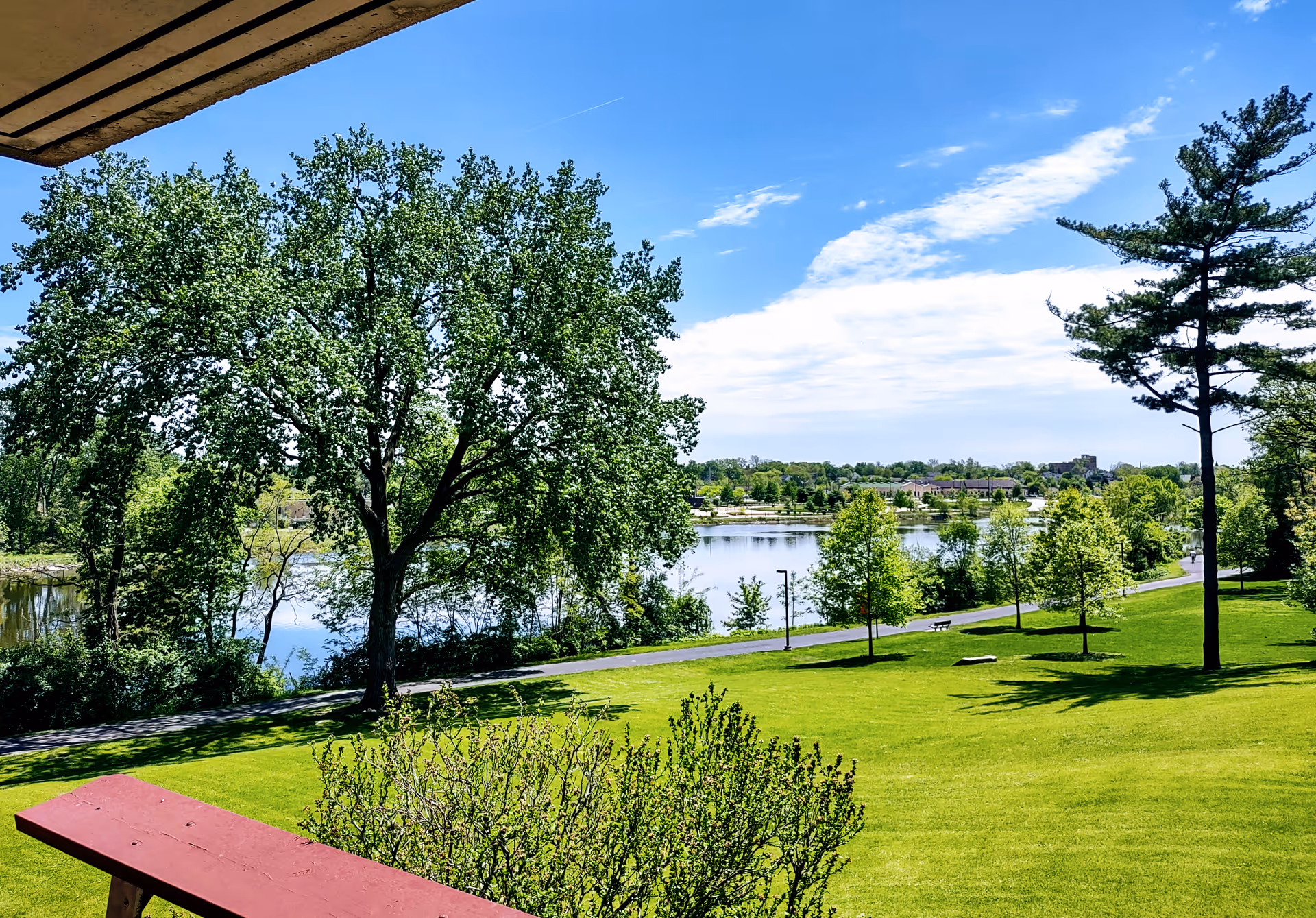View of a lush green lawn with several trees and a walking path next to a calm river under a bright blue sky with some clouds, seen from a covered porch with a red railing.