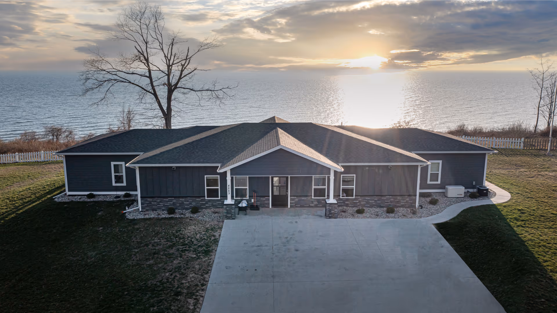 Front exterior view of a single-story building with a dark roof and gray siding, situated near a large body of water with a sunset in the background. There is a concrete driveway leading to the entrance, a large leafless tree to the left, and a white picket fence along the water's edge.