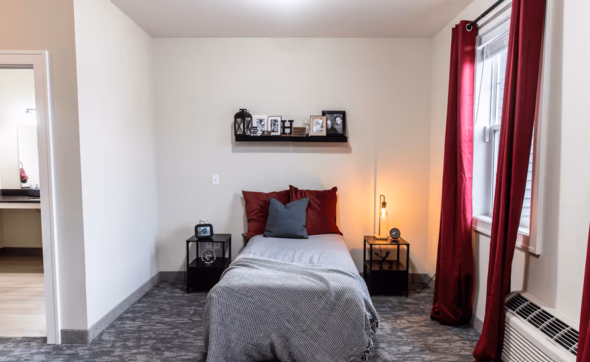 A small, neatly arranged bedroom with a single bed centered against a white wall. The bed has a gray blanket and red and gray pillows. On either side of the bed are black nightstands, one with a small clock and the other with a lamp and decorative items. Above the bed is a black floating shelf with framed photos and decorative pieces. To the right, a window with red curtains lets in natural light. To the left, an open doorway leads to a bathroom area.