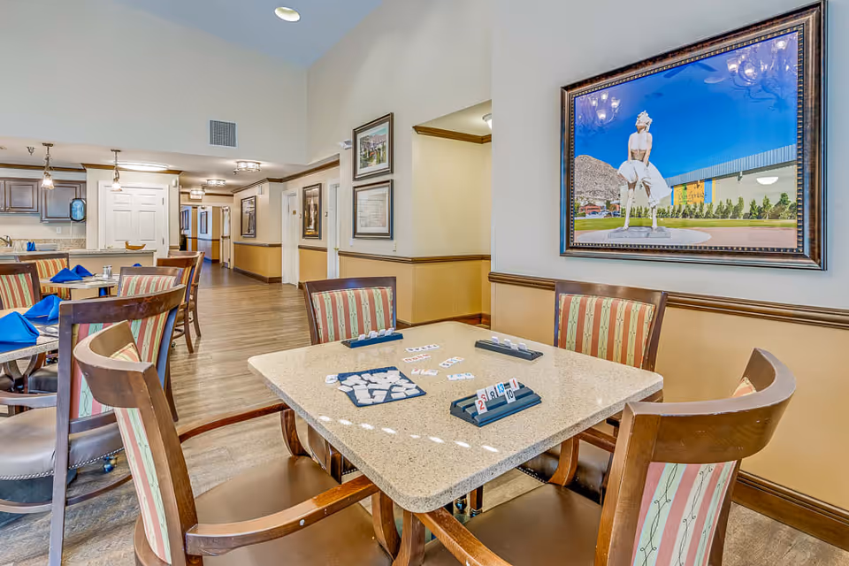 Interior view of a common area in a senior living facility with a table set up for a tile-based game, surrounded by wooden chairs with striped cushions. In the background, there are more tables with blue napkins, a kitchen area, and framed artwork on the walls, including a large framed photo of a statue outdoors.