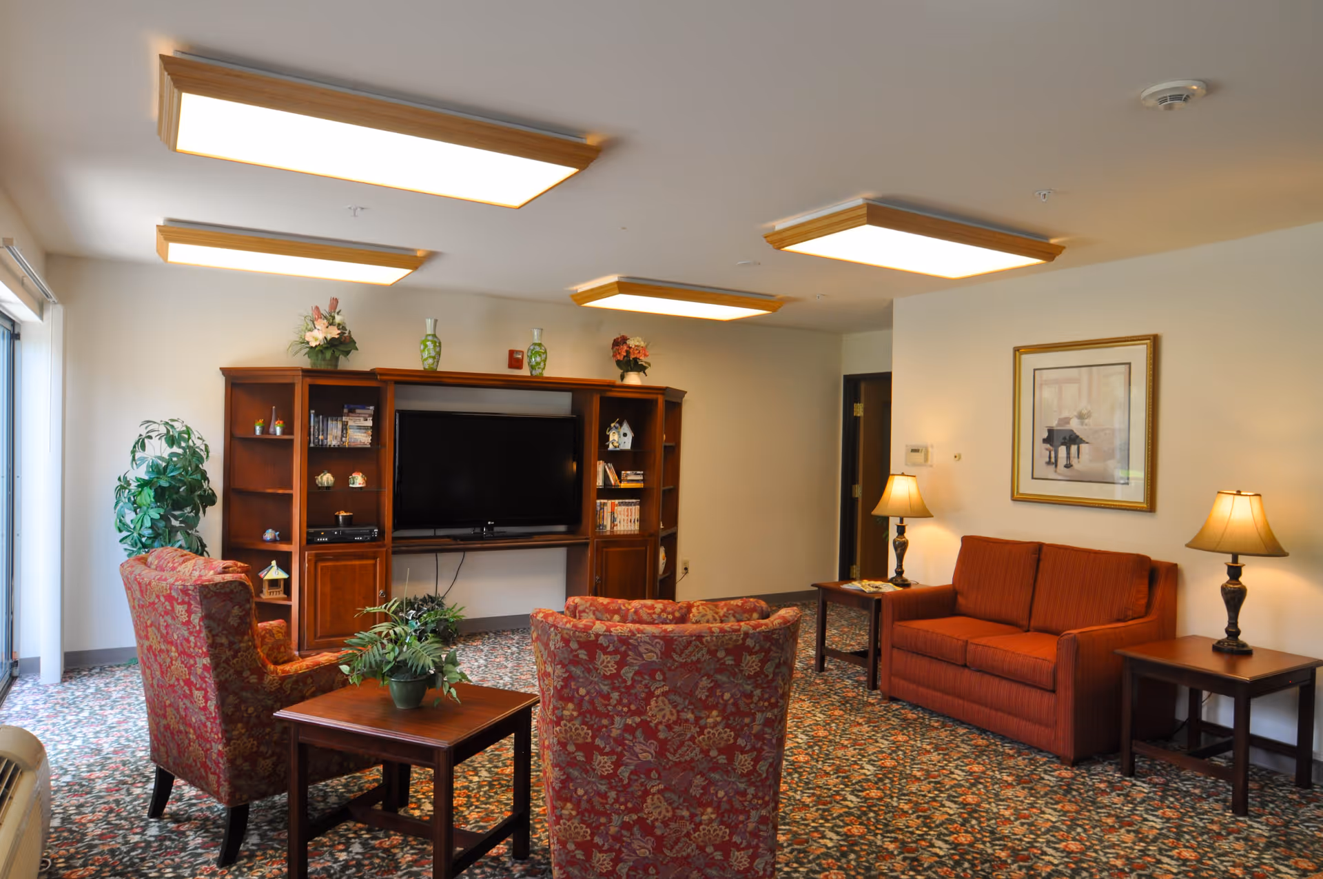 A cozy living room area in a senior living facility featuring a red patterned armchair and a matching sofa, a wooden coffee table with a potted plant, a large wooden entertainment center with a flat-screen TV, decorative vases, and shelves with books and ornaments. The room has floral carpet flooring, soft lighting from ceiling fixtures and table lamps, and a framed picture on the wall.