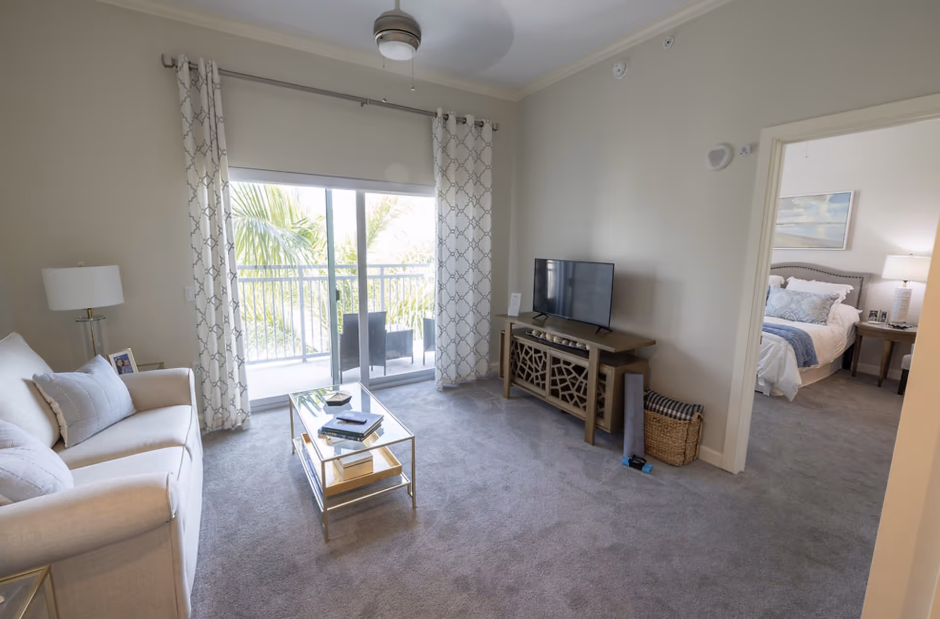 Bright living room with sliding glass doors to a balcony, a sofa, coffee table, TV on a console, and a view into an adjacent bedroom.