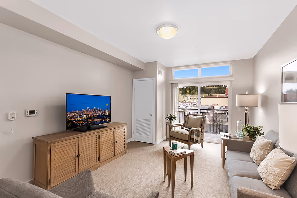 Bright furnished living room with a sofa, armchair, TV on a wooden console, and a sliding glass door leading to a balcony.