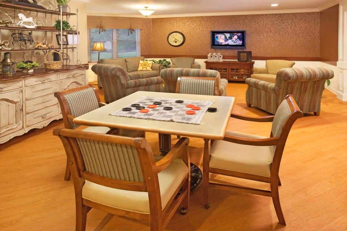 Cozy common room with a game table and chairs in the foreground and sofas, armchairs and a wall-mounted TV in the background.