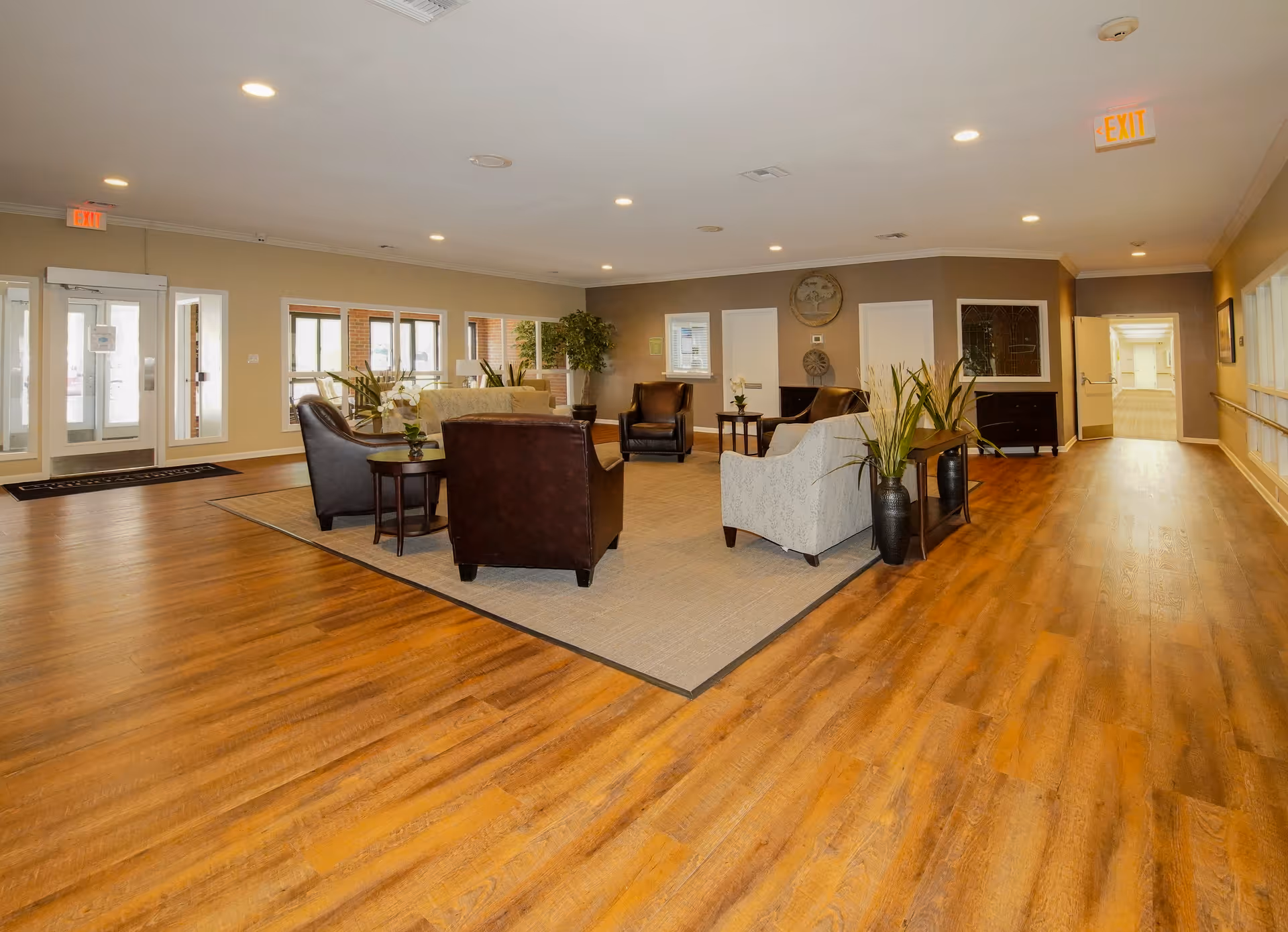 Spacious senior living facility lounge with wood floors, a seating area of sofas and armchairs, potted plants, and entry doors.