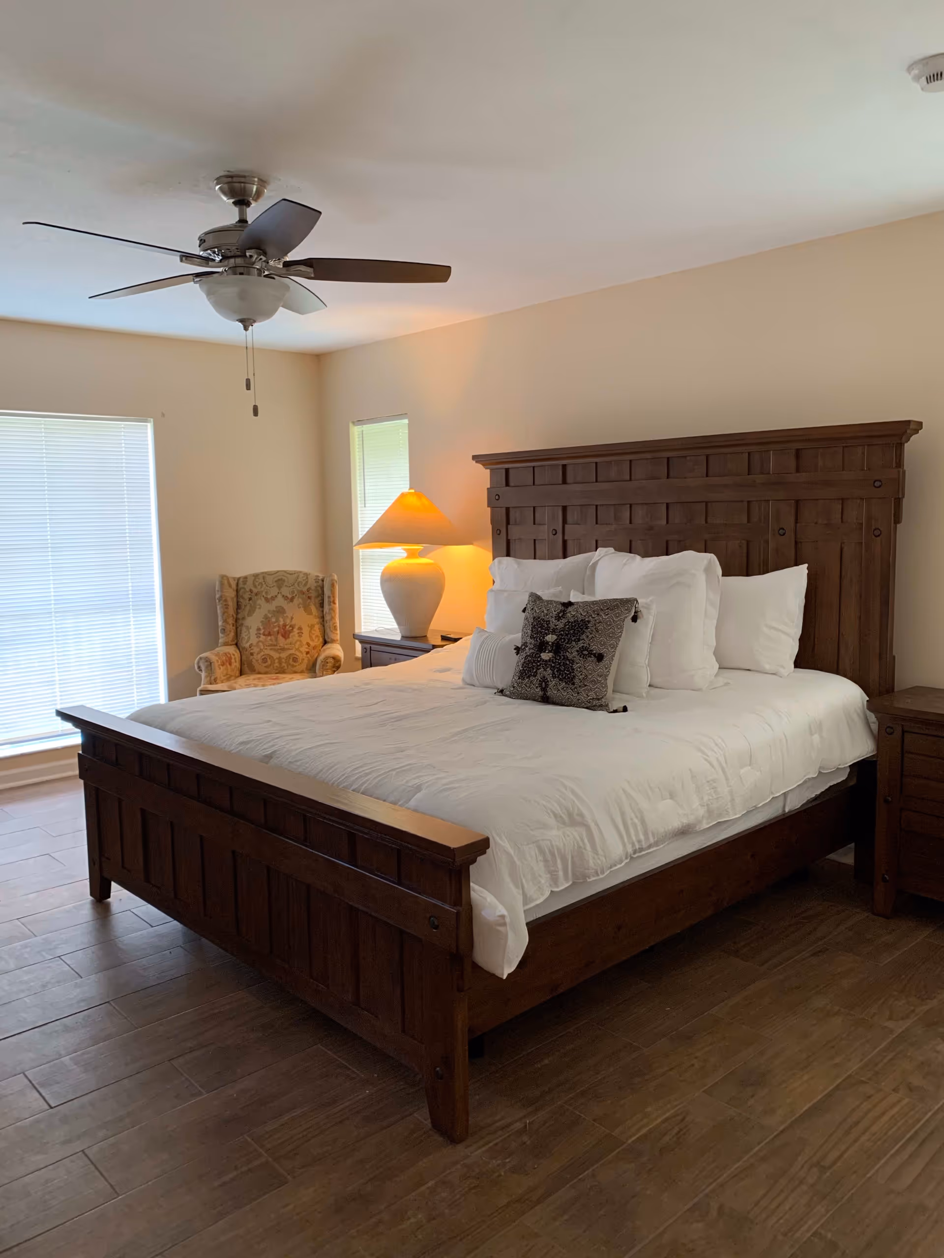 A bedroom with a large wooden bed frame and white bedding, several pillows, a wooden nightstand with a lamp, a floral upholstered armchair, two windows with blinds, and a ceiling fan.