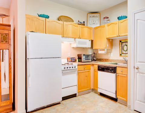 A compact kitchen with light wood cabinets, a white refrigerator, white stove, microwave, dishwasher, and a toaster oven on the countertop. The kitchen has a beige countertop and a tiled floor. Decorative items are placed on top of the cabinets and a framed picture hangs on the wall.