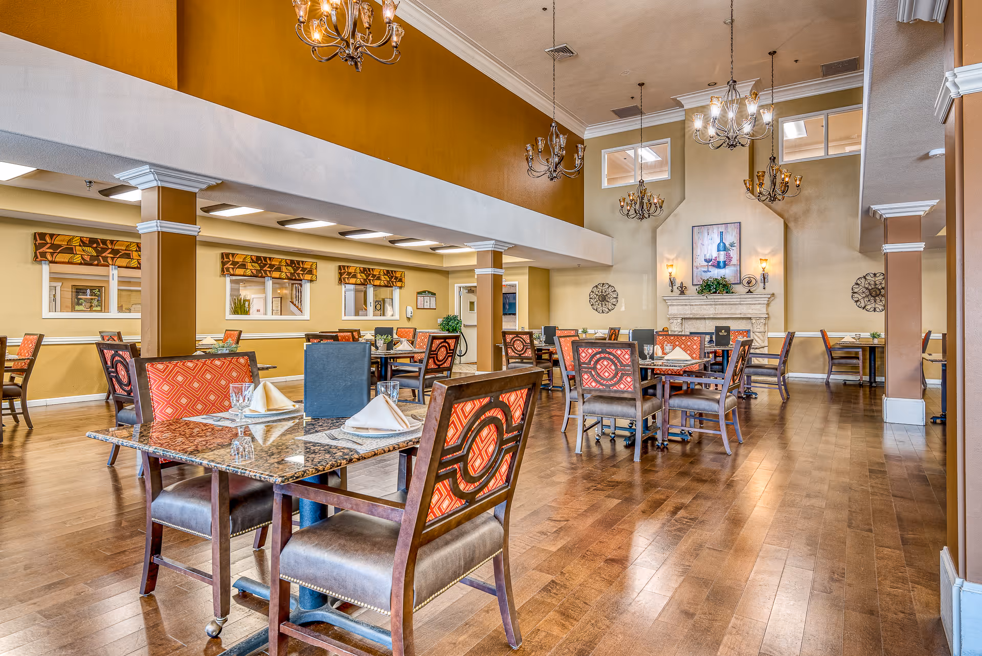 A spacious dining room with multiple tables and chairs arranged neatly. The chairs have patterned red upholstery and the tables are set with napkins and glassware. The room features wooden flooring, warm yellow and beige walls, decorative chandeliers hanging from a high ceiling, and a fireplace with artwork above it.