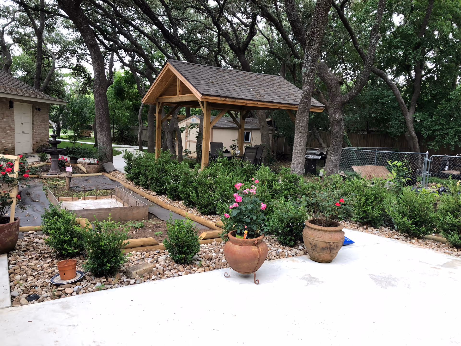 Outdoor garden area with a wooden gazebo, surrounded by trees and various potted plants including roses. There is a stone pathway, a small fenced area, and a brick building visible on the left side.