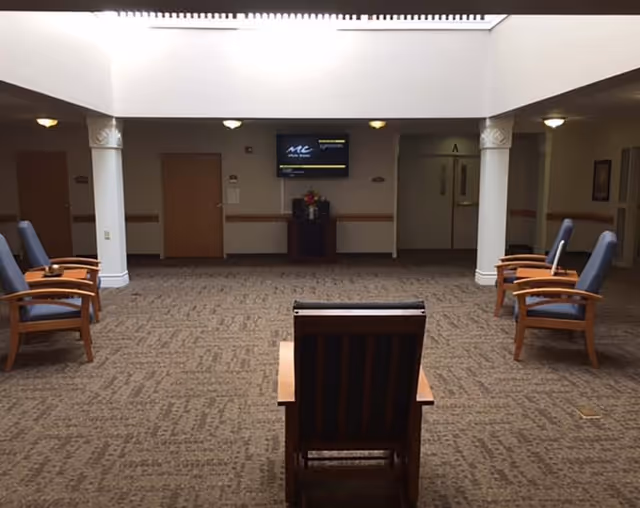 A spacious interior common area with carpeted floor, several wooden chairs with blue cushions arranged along the sides, two white columns, and a wall-mounted television screen displaying a message. The area is well-lit with ceiling lights and has multiple closed doors along the back wall.