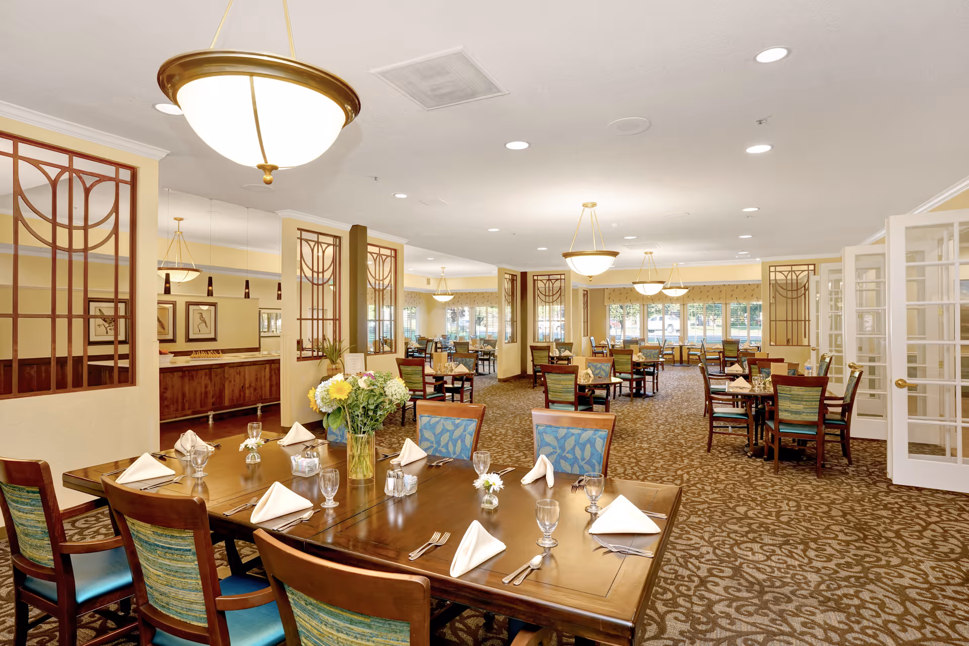 A spacious and well-lit dining room with multiple wooden tables and chairs arranged neatly. Each table is set with folded white napkins, glassware, and silverware. There are decorative light fixtures hanging from the ceiling and large windows along the far wall allowing natural light to fill the room. The carpet has a patterned design, and there are decorative wooden panels and mirrors on the walls.