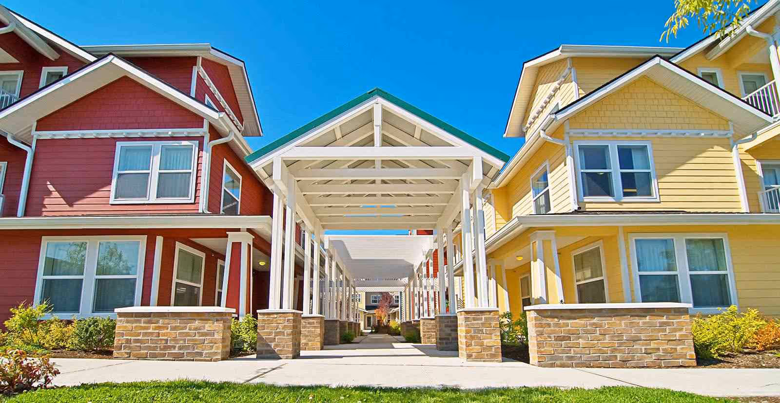Covered entrance walkway between two colorful multi-story residential buildings with brick planters and a green lawn under a clear blue sky.