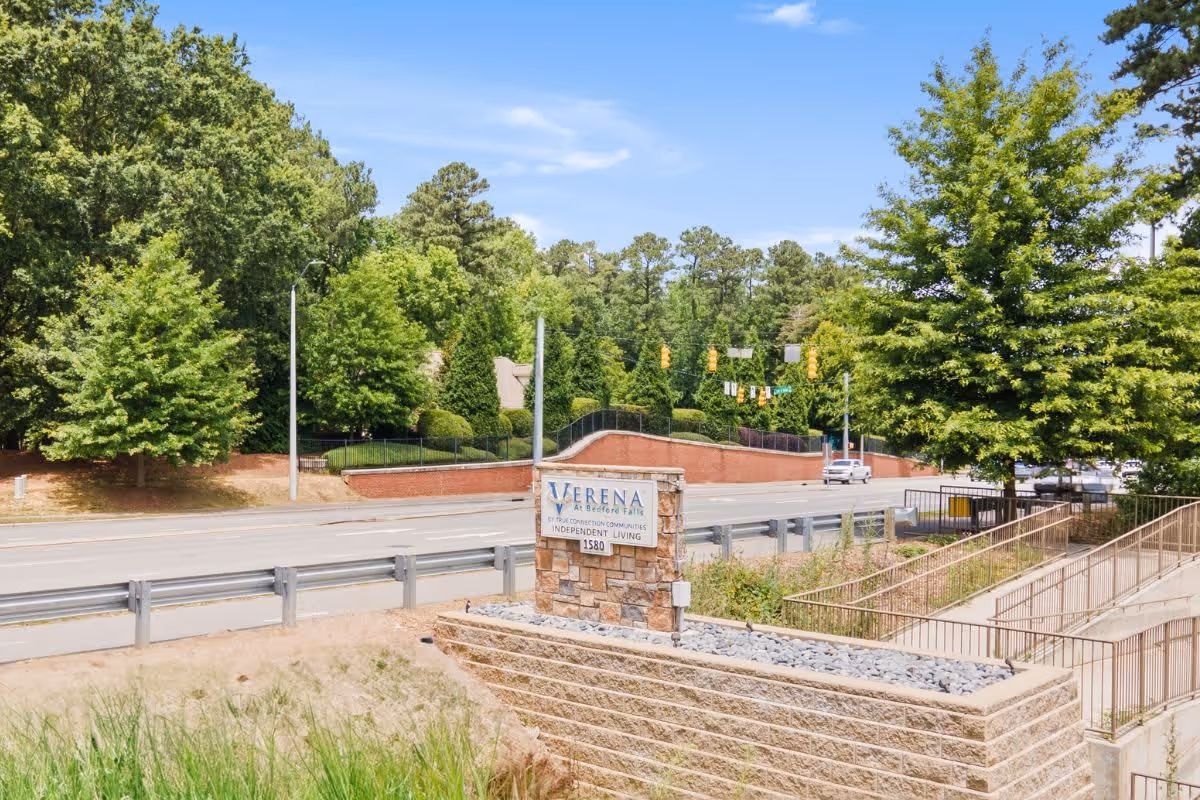 Outdoor view of the entrance sign for Verena at Bedford Falls independent living community, located beside a road with trees and greenery in the background under a blue sky.