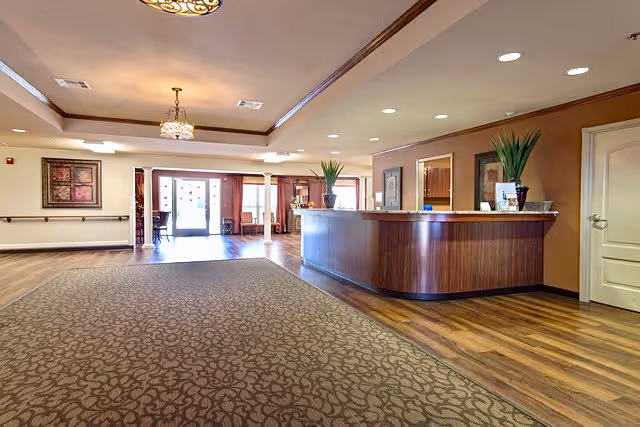 Spacious and well-lit reception area with a wooden front desk, decorative plants on the counter, patterned carpet, wooden flooring, and framed artwork on the walls. Large windows and glass doors allow natural light to fill the space.