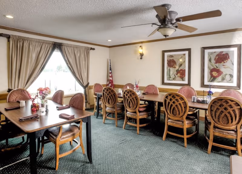 A dining room with two wooden tables and multiple chairs arranged around them. The tables have napkins and small flower arrangements. There are two framed floral paintings on the wall, a window with beige curtains, a ceiling fan, and an American flag in the corner.