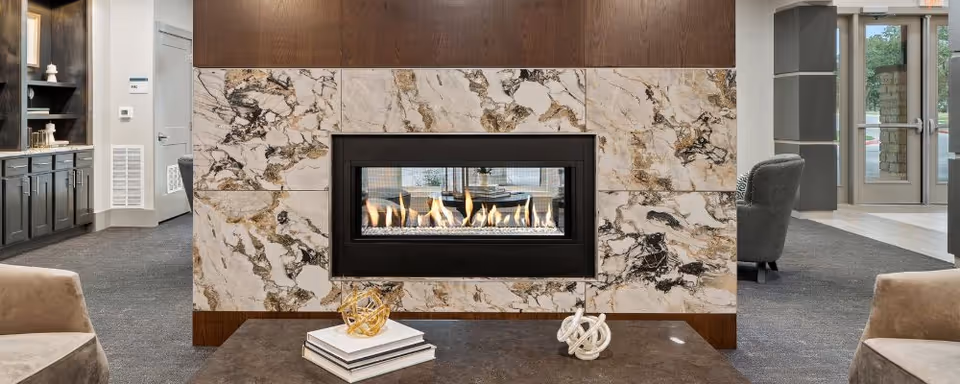 A modern interior lounge area featuring a rectangular gas fireplace set in a marble and wood-paneled wall. In front of the fireplace is a coffee table with decorative objects and books. The room has carpeted flooring, comfortable armchairs, and large glass doors leading outside.