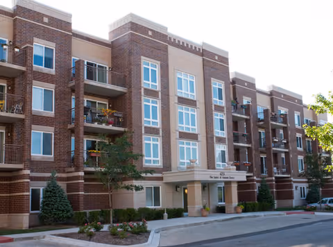 Exterior view of a multi-story senior living facility building with brick and beige facade, balconies, windows, small trees, and landscaped bushes near the entrance.
