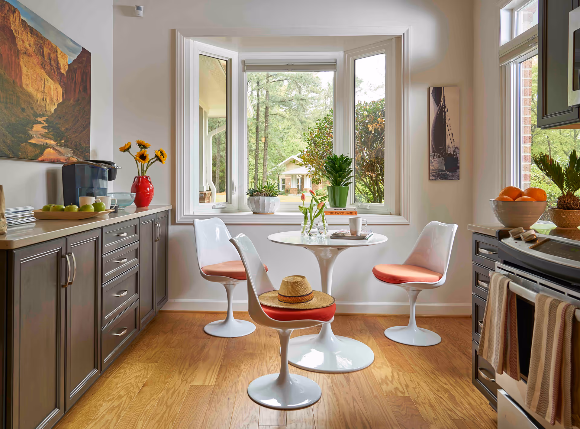 Sunlit kitchen dining nook with a small round table, three modern white chairs with orange cushions, cabinets and a stove, and a bay window looking out to greenery.