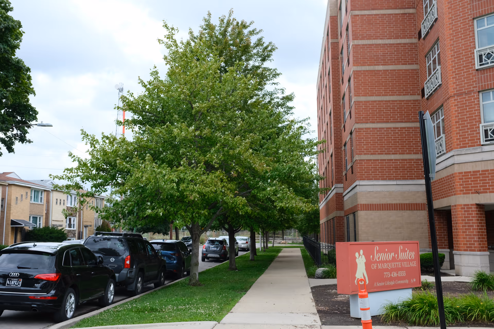 Sidewalk lined with green trees and parked cars next to a multi-story red brick building with a sign that reads Senior Suites of Marquette Village, a senior living community.