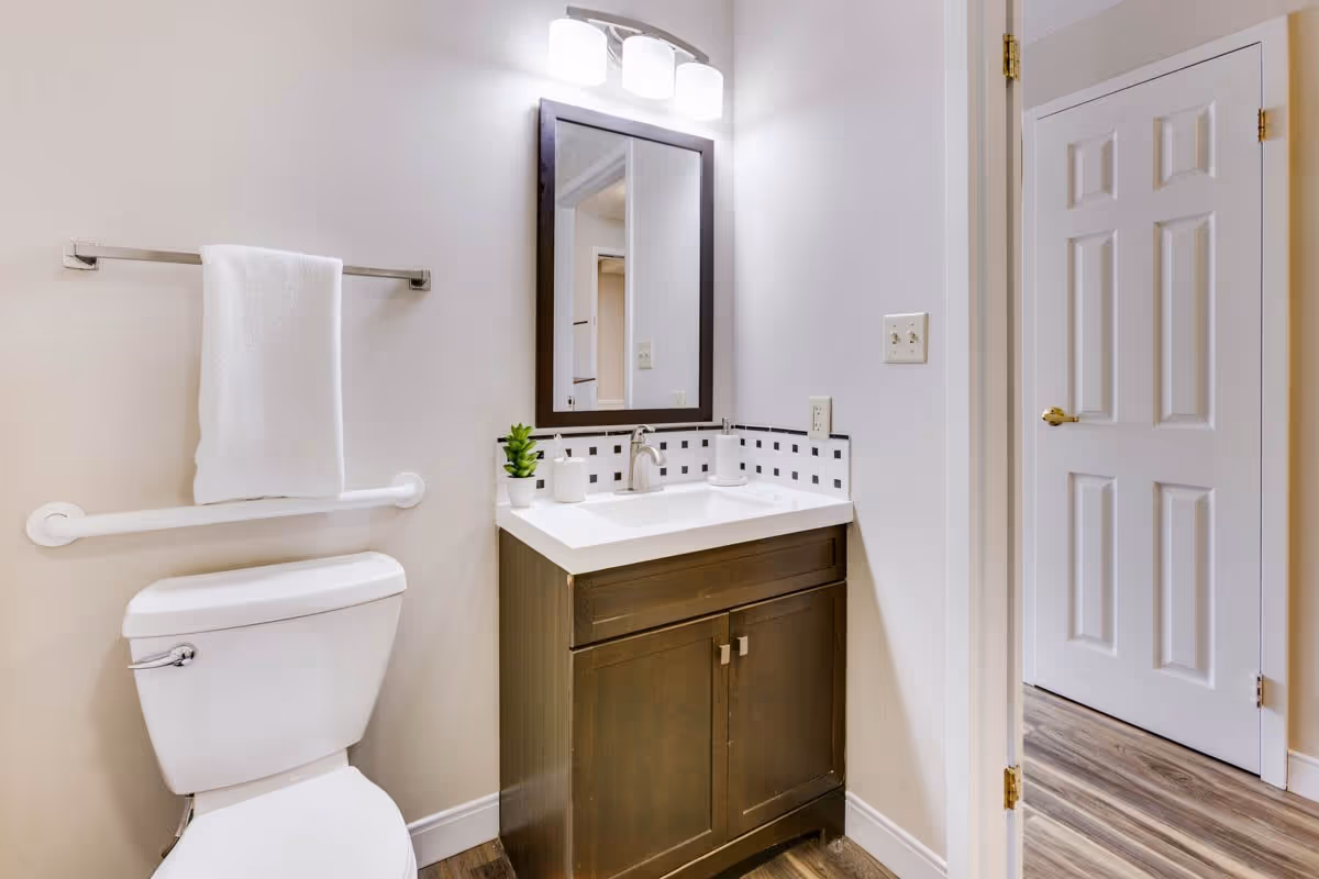 A clean bathroom featuring a white toilet with a towel hanging on a metal bar above it, a dark wood vanity with a white countertop, a rectangular mirror with a dark frame above the sink, and a small green plant and soap dispensers on the countertop. The bathroom has light-colored walls and wood-look flooring, with a white door visible to the right.