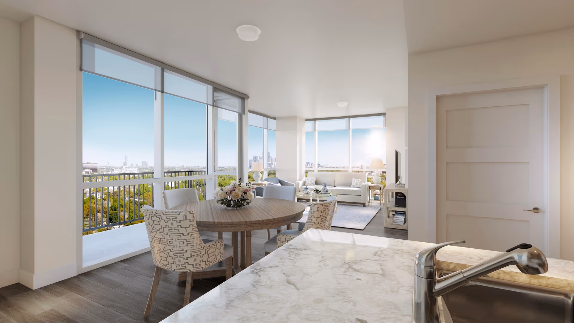 Bright and modern open-concept living space with large floor-to-ceiling windows offering a panoramic city view. The room features a round wooden dining table with four patterned chairs, a cozy sitting area with a sofa, armchairs, a coffee table, and two lamps. A marble countertop with a sink and faucet is visible in the foreground, and a white door is on the right side of the room.