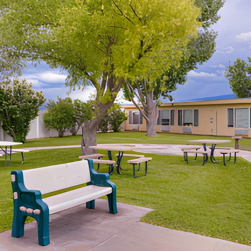 Outdoor courtyard with a green-and-white bench, picnic tables on grass, trees and a single-story building in the background.