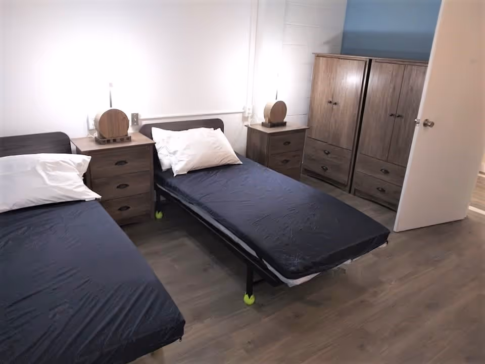 A simple bedroom with two single beds featuring dark blue fitted sheets and white pillows. Between the beds is a wooden nightstand with a round decorative lamp. There are two wooden wardrobes against the wall and a door partially open leading to another room. The floor is wooden and the walls are white.
