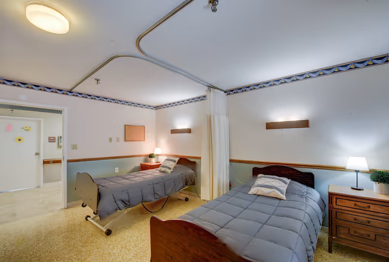 A senior living facility bedroom with two single beds separated by a privacy curtain. Each bed has a gray comforter and a pillow. There are two wooden nightstands with lamps and a small plant on one. The room has a light fixture on the ceiling and a door leading to a hallway.
