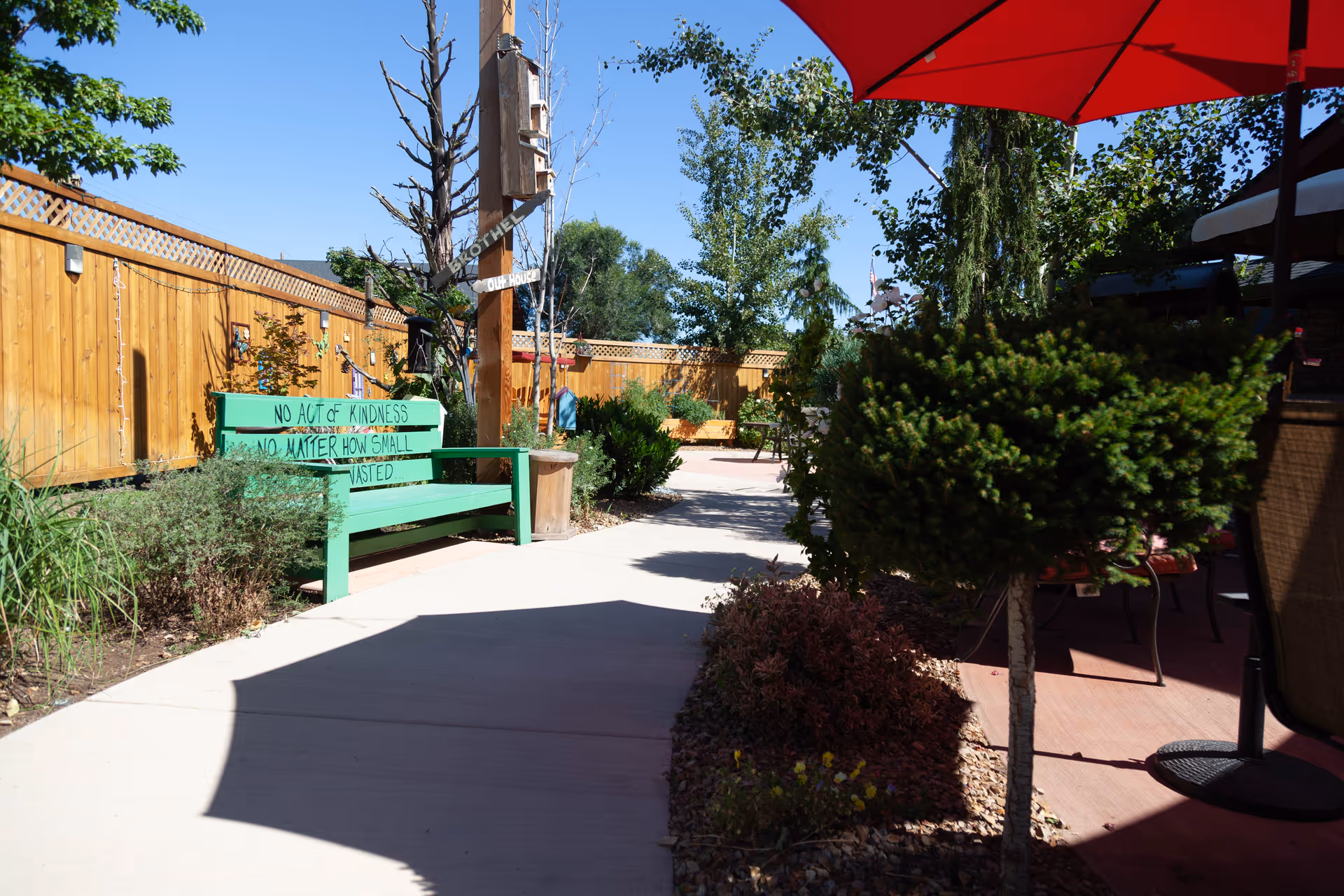 Outdoor garden area with a concrete pathway, green bench with the inscription 'No act of kindness no matter how small is wasted,' surrounded by plants and trees, wooden fence in the background, and a red umbrella providing shade over a seating area.