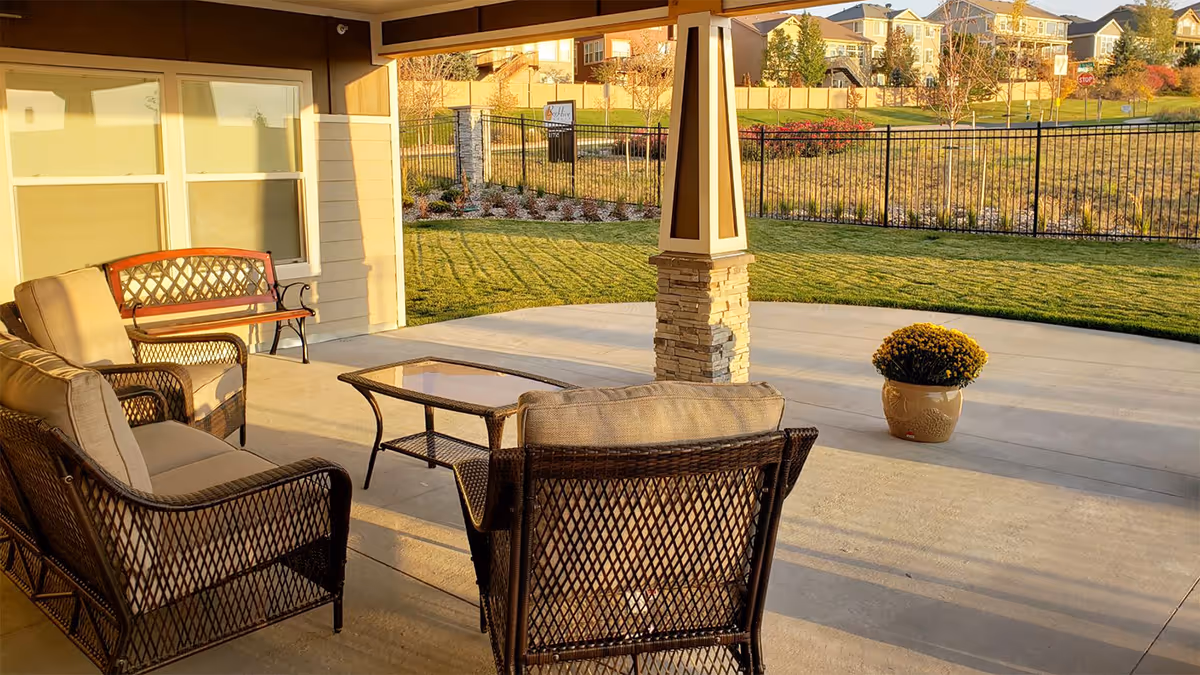 Covered outdoor patio area with cushioned wicker chairs, a glass-top coffee table, and a wooden bench. The patio overlooks a fenced grassy yard with houses and trees in the background, illuminated by warm sunlight.