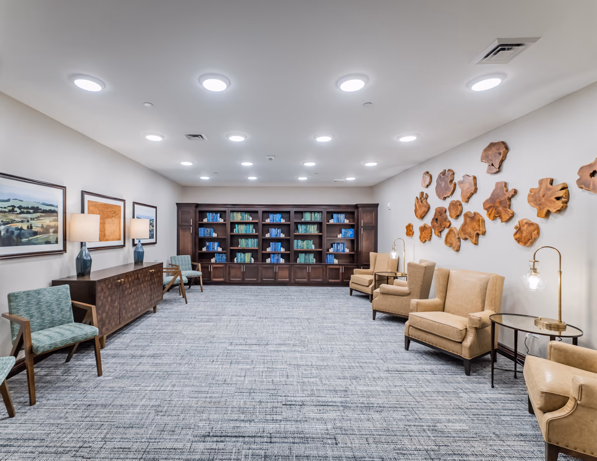 A spacious, well-lit senior living common area with a large bookshelf filled with books at the far end. The room features beige armchairs with side tables and lamps on the right side, green patterned chairs on the left, and a wooden sideboard with two lamps and framed artwork above it. The wall on the right is decorated with wooden art pieces resembling tree slices.