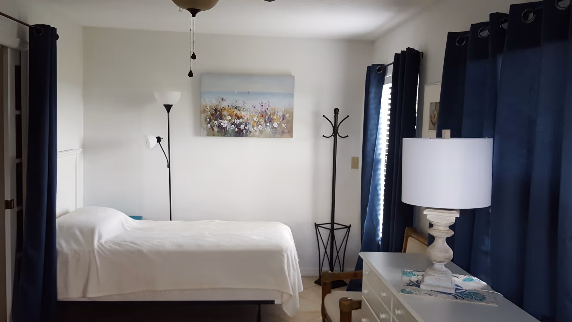 A bright bedroom with a single bed covered in white linens, blue curtains, a dresser with a lamp, and wall art above the bed.