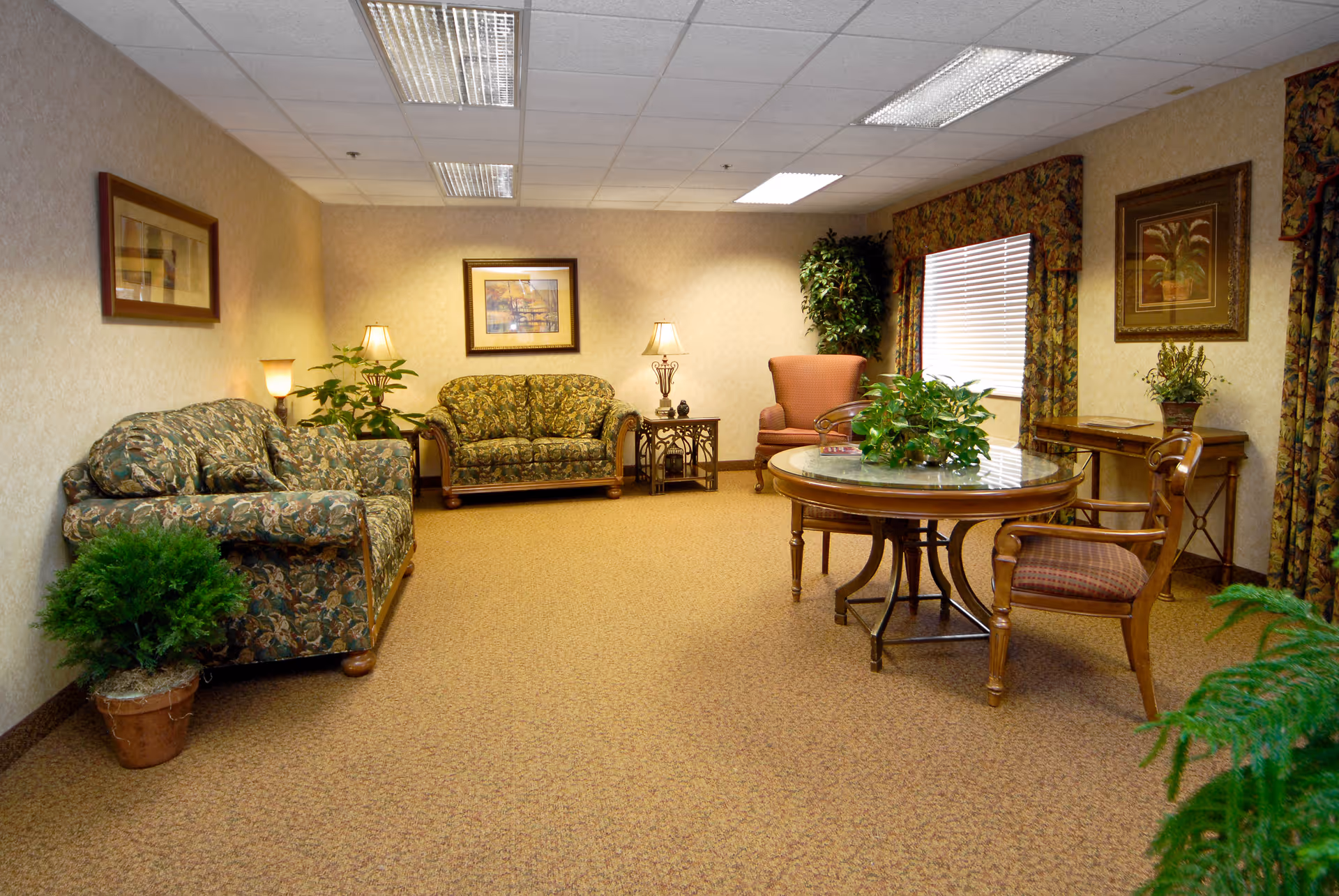 A cozy living room area with floral patterned sofas, a round wooden table with chairs, potted plants, framed artwork on the walls, and warm lighting from table lamps. The room has beige walls and carpet, with a window covered by floral curtains.