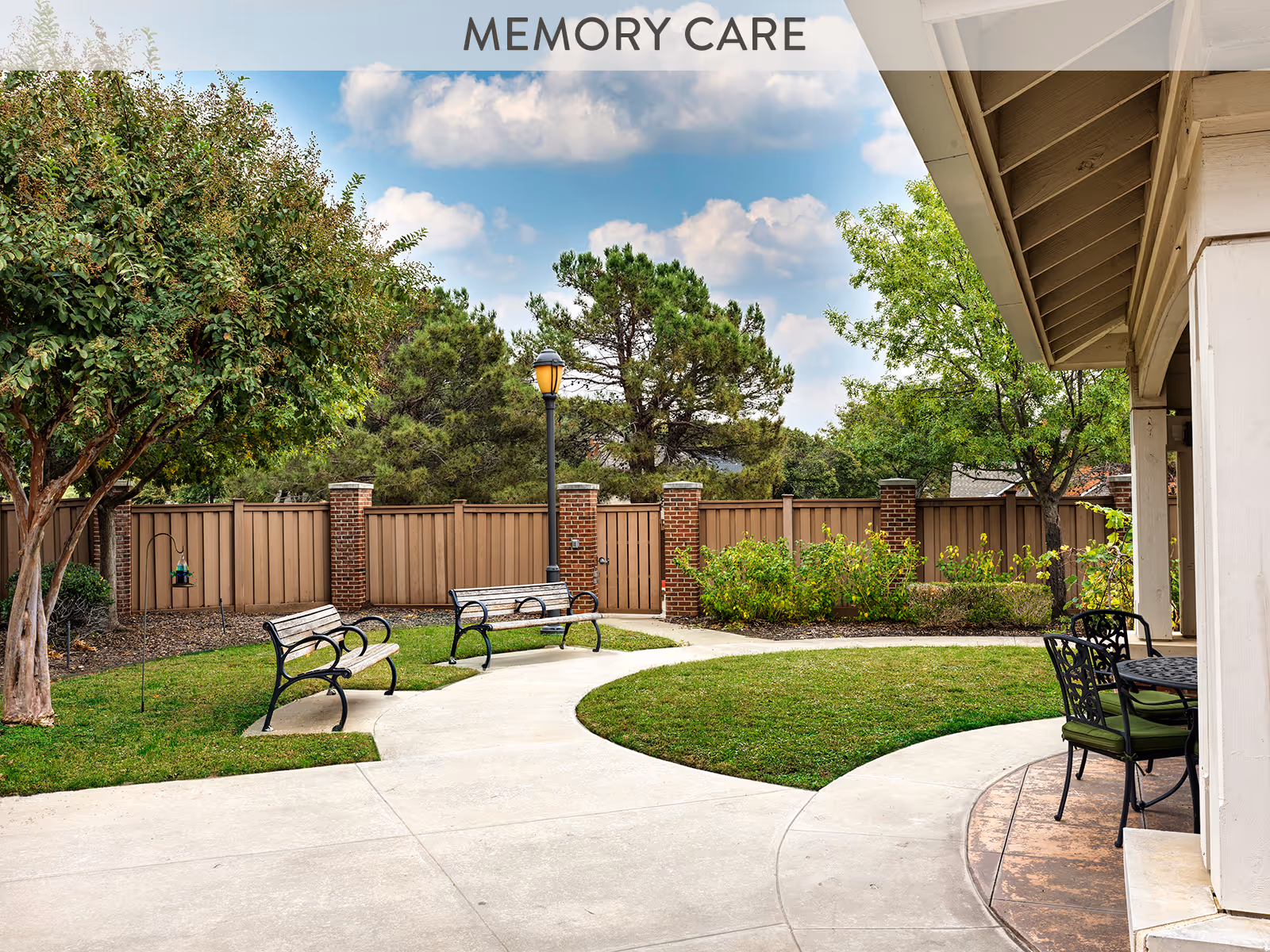 Outdoor courtyard area with a curved concrete pathway, two wooden benches with black metal frames, a lamppost, green grass, trees, and a wooden fence with brick pillars. There is also a covered patio area with a round table and chairs with green cushions.