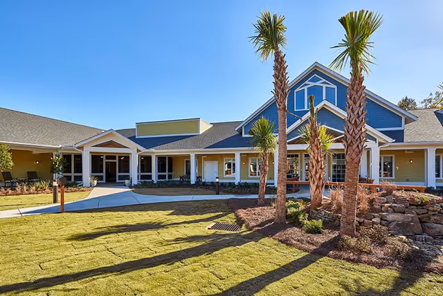 Exterior view of a senior living facility building with a well-maintained lawn, palm trees, and a clear blue sky. The building features a combination of yellow and blue siding with white trim and a covered entrance walkway.