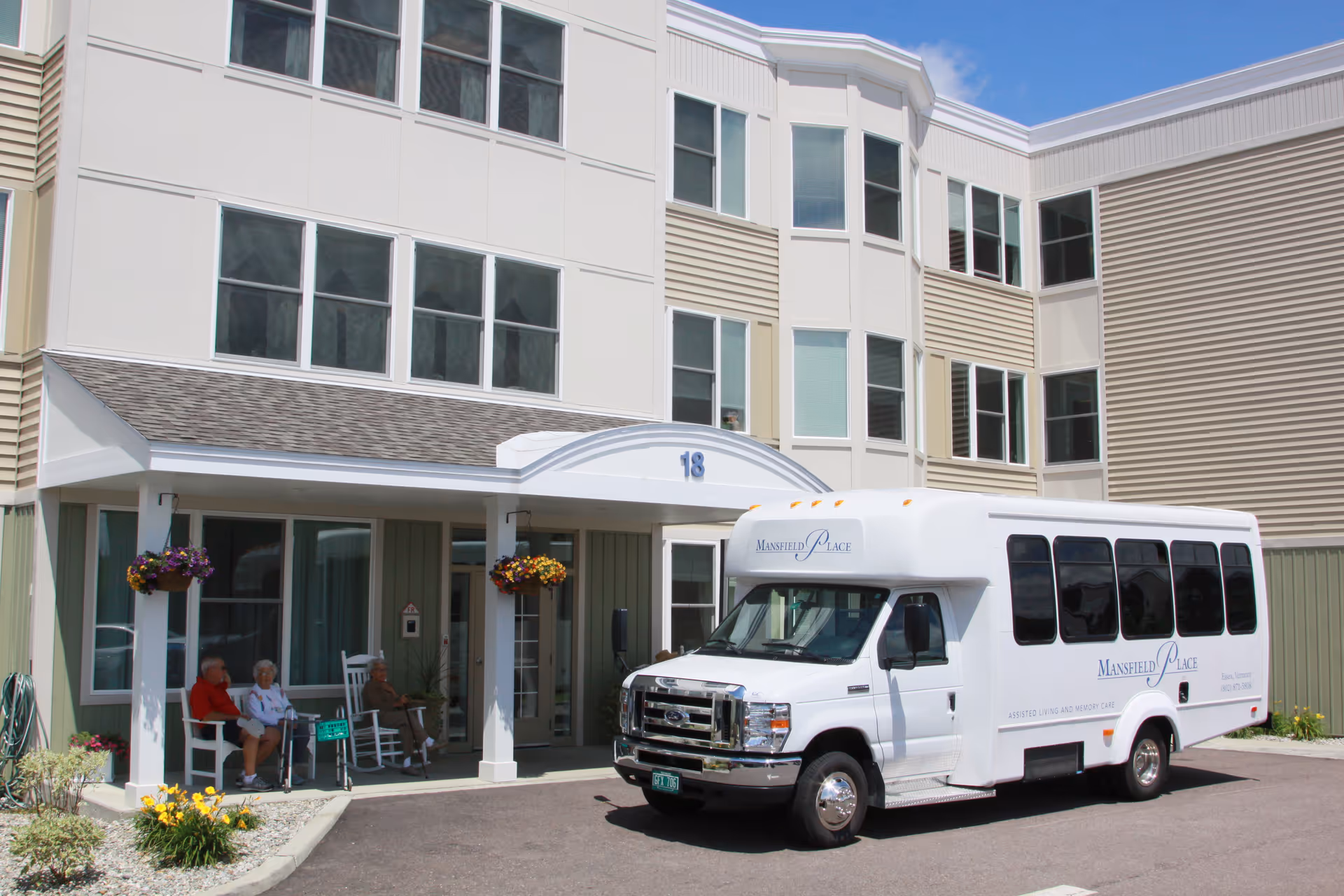 Front entrance of Mansfield Place senior living facility with a white shuttle bus parked and residents seated on the covered porch.