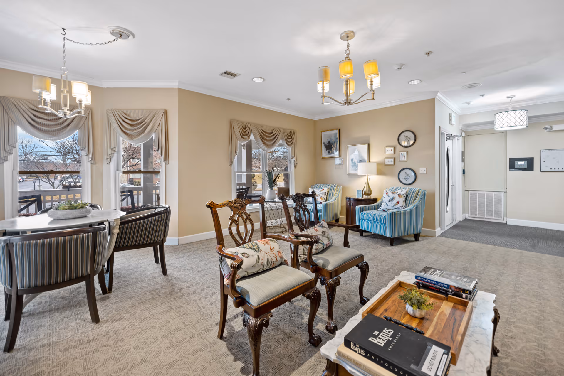 A cozy living room area in a senior living facility with two wooden armchairs featuring floral cushions, two striped upholstered armchairs, a coffee table with books and a small plant, and a dining table with four striped chairs near windows with beige drapes. The walls are painted beige and decorated with framed pictures and clocks. The room is well-lit with ceiling lights and natural light from the windows.