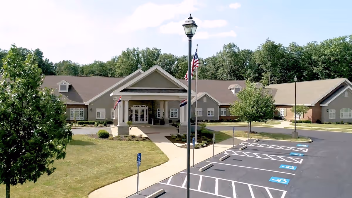 Exterior view of a single-story senior living facility building with a covered entrance, surrounded by trees and a parking lot with designated handicap parking spaces.
