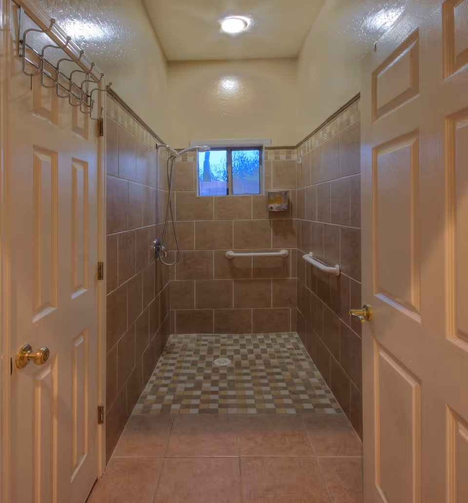 A tiled walk-in shower with beige and brown square tiles on the floor and walls, two white grab bars on the side walls, a small window above, a mounted soap dispenser, and a handheld showerhead. The shower area is framed by two open doors with gold-colored handles and a metal hook rack on the left door.