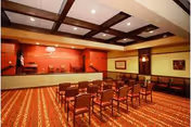 A large meeting or event room with rows of chairs facing a raised stage area. The room has a patterned carpet, a coffered ceiling with recessed lighting, and red walls behind the stage. There are additional chairs lined up against the walls and framed artwork on the walls.