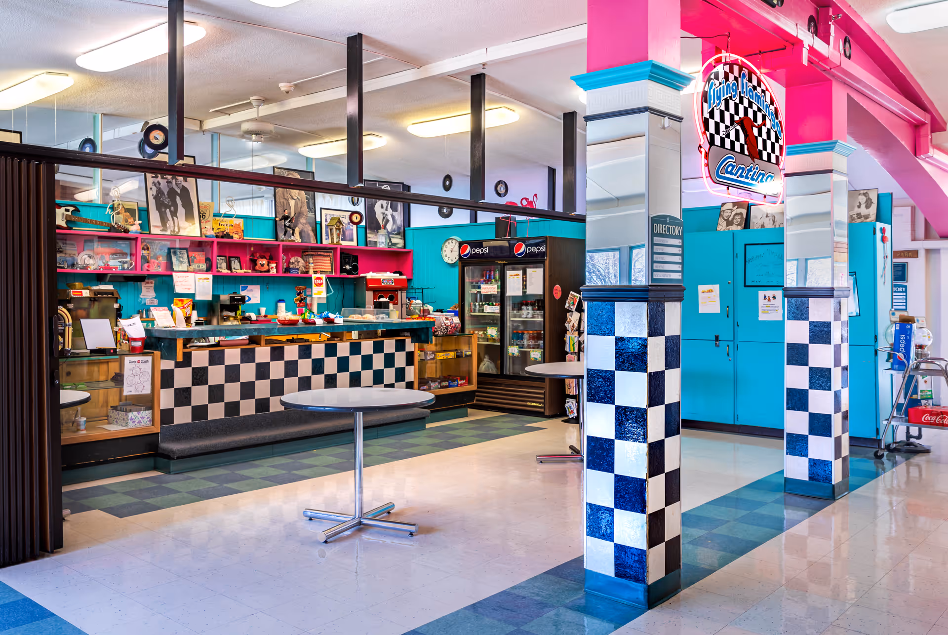 Retro-themed dining/cafeteria area with checkered columns, a service counter, neon sign, refrigerators, and round tables.