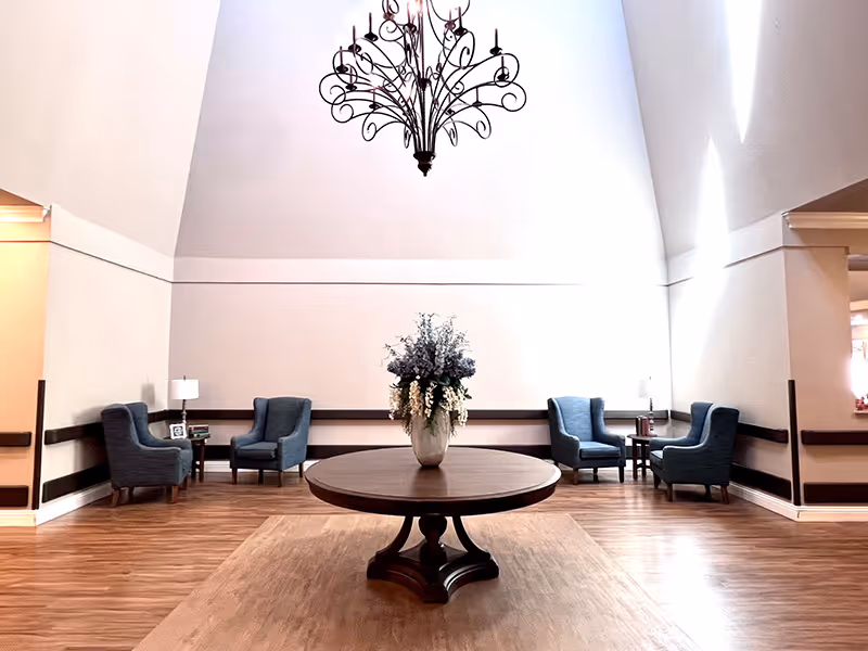 Bright lobby-style sitting area with a round wooden table and floral centerpiece under a chandelier, flanked by blue armchairs.