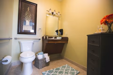 A clean bathroom with a white toilet, a wicker basket holding towels, a wall-mounted sink with a mirror above it, a framed decorative picture on the wall, a patterned rug on the tiled floor, and a dark wooden cabinet with a vase of orange flowers on top.