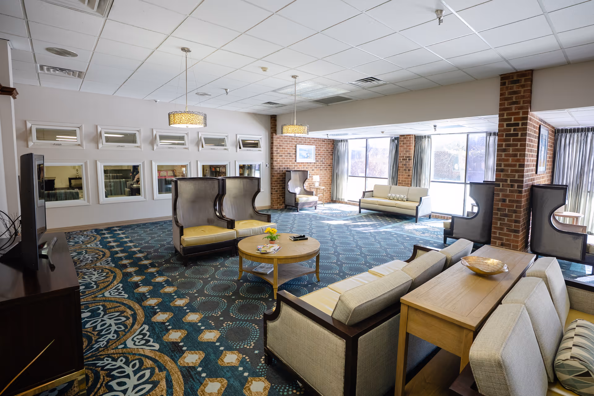 A spacious senior living facility common area with patterned carpet, multiple seating arrangements including sofas and high-backed chairs, large windows letting in natural light, brick pillars, and pendant lights hanging from a white tiled ceiling.
