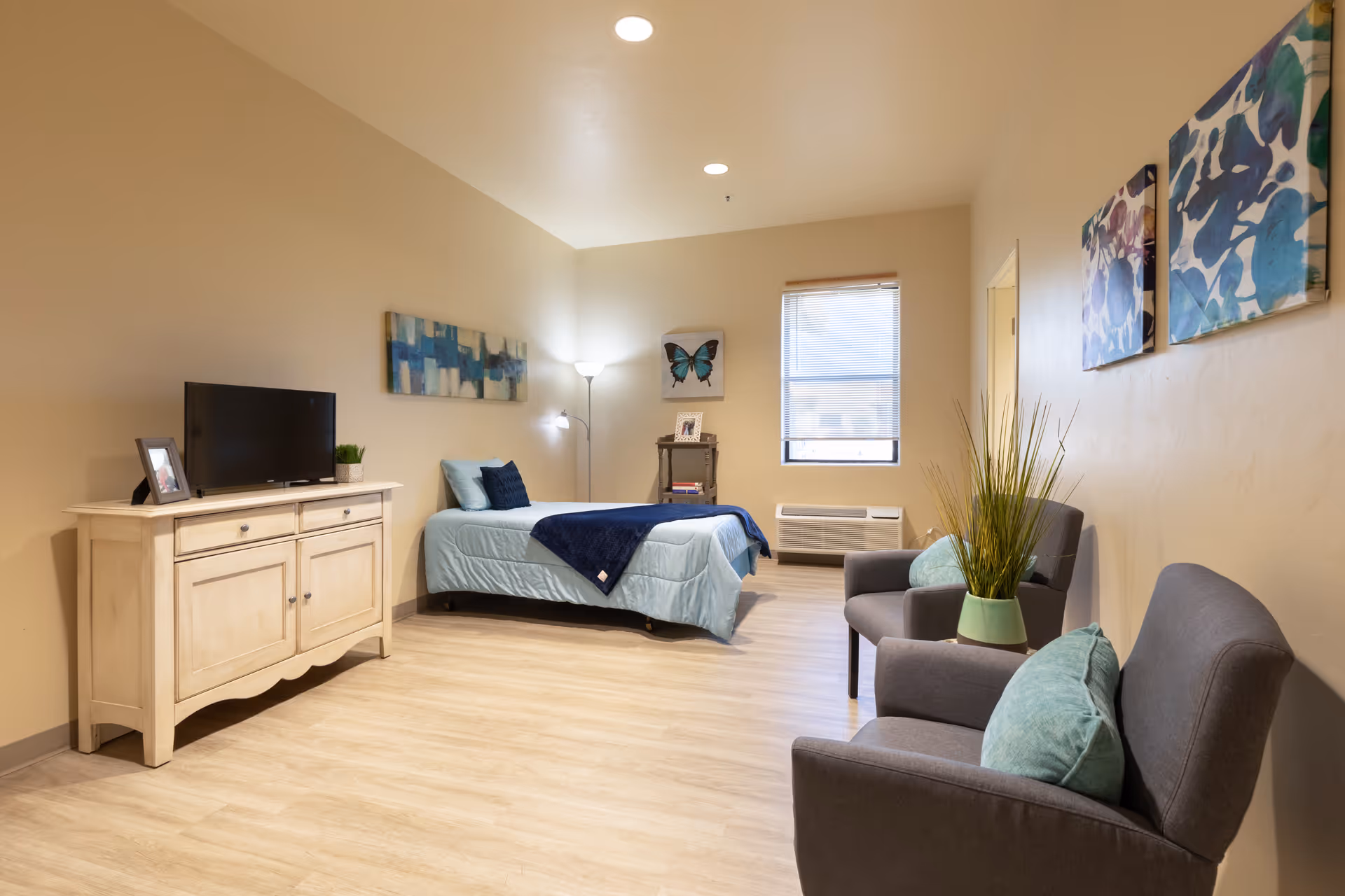 A bright and cozy assisted living bedroom featuring a single bed with light blue bedding and a dark blue throw blanket. The room includes a wooden dresser with a TV and a small plant, two gray armchairs with teal cushions, a potted plant on a small table between the chairs, and colorful artwork on the walls. A window with blinds allows natural light to enter the room.