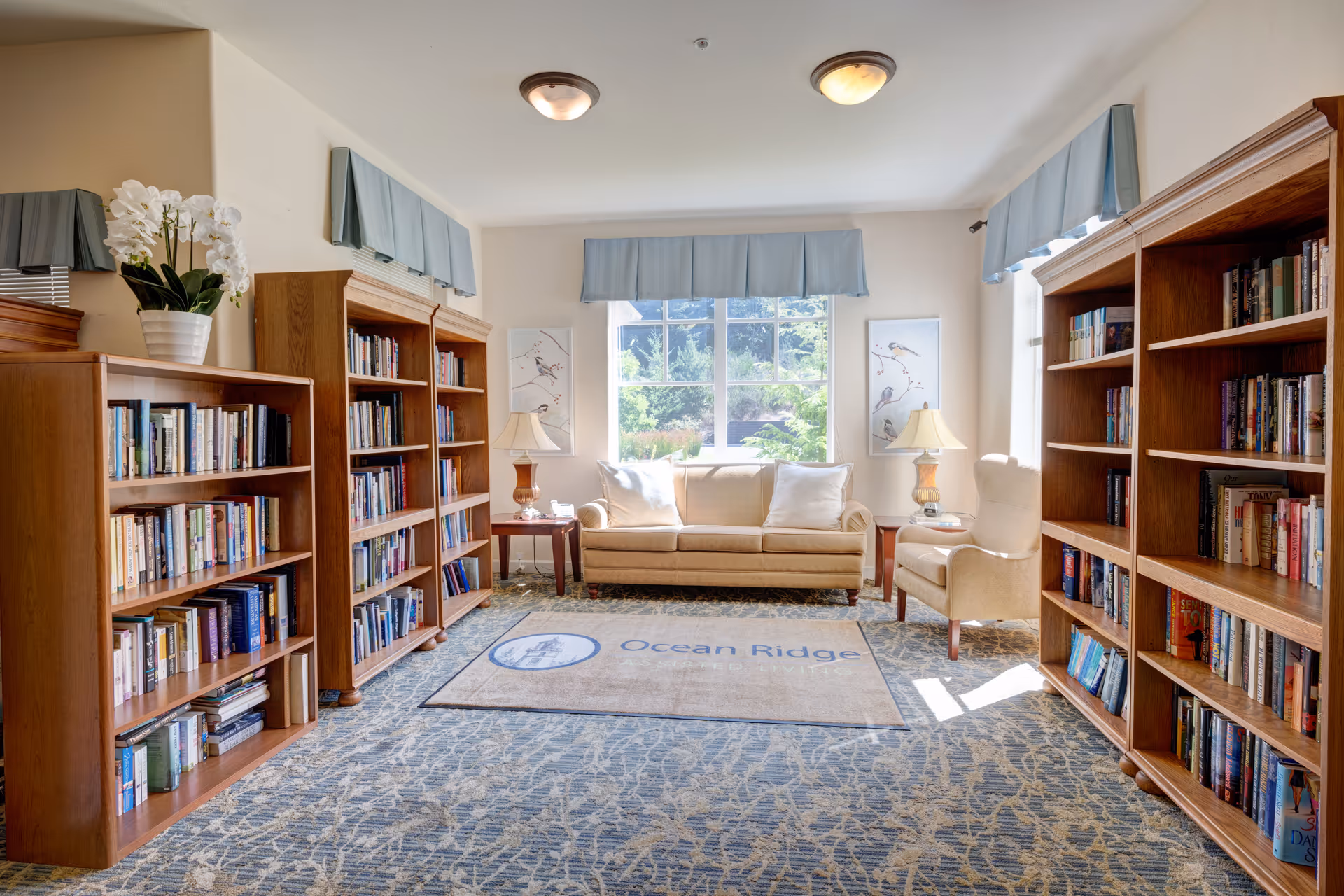 A bright and cozy library room with large windows letting in natural light. The room features wooden bookshelves filled with books on both sides, a beige sofa with white pillows, a beige armchair, two side tables with lamps, and a carpet with the Ocean Ridge logo. Light blue valances adorn the windows.