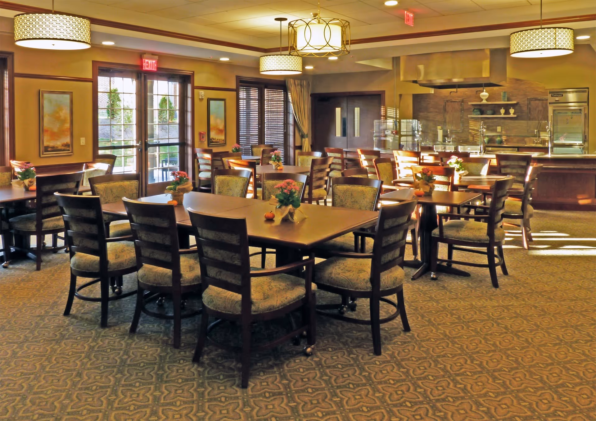 A well-lit dining room with multiple wooden tables and cushioned chairs arranged neatly. Each table has a small floral centerpiece. The room features large windows with wooden blinds, framed artwork on the walls, and modern ceiling light fixtures. In the background, there is a kitchen area with stainless steel appliances and a serving counter.