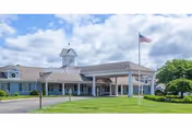 Front entrance of a single-story senior living facility with a porte-cochère, American flag on a flagpole, and a manicured lawn under a blue sky.