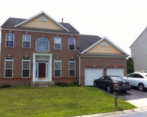 Two-story brick house with an attached two-car garage, front lawn, and parked cars.